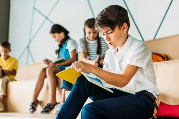 Joven colegial escribiendo en cuaderno mientras sus compañeros de clase se sientan detrás - foto de stock