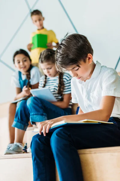Jeune écolier écrit dans un cahier tandis que ses camarades de classe assis derrière — Photo de stock