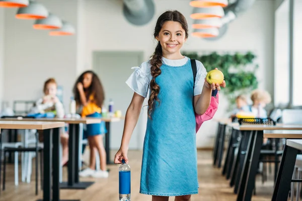 Усміхнена маленька школярка з яблуком і пляшкою води в шкільних кафетеріях — стокове фото