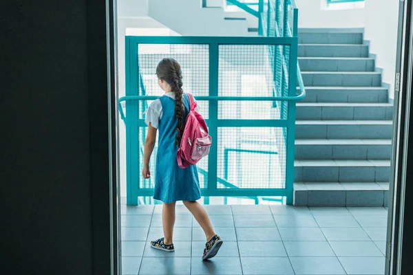 Vista trasera de la colegiala en vestido caminando en el pasillo de la escuela con mochila - foto de stock