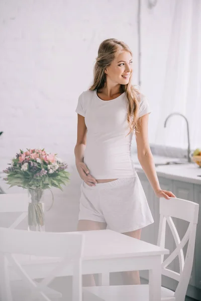 Donna incinta sorridente in piedi a tavola con mazzo di fiori in vaso a casa — Foto stock