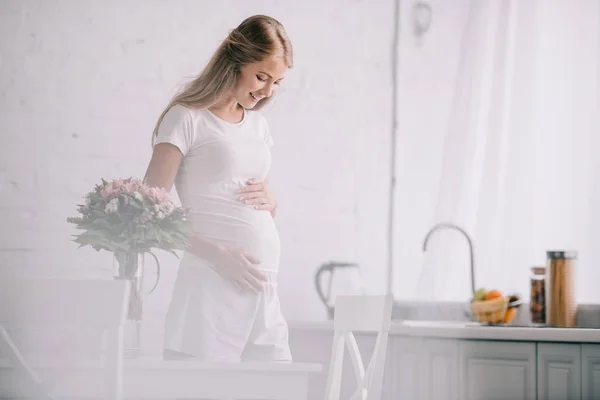 Happy pregnant woman standing at table with bouquet of flowers in vase at home — Stock Photo