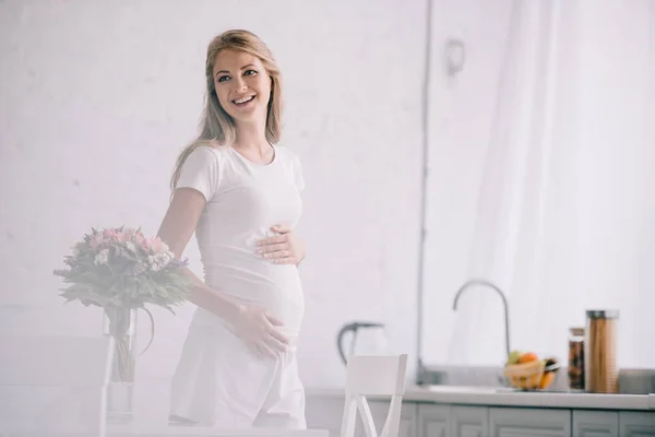 Portrait of happy pregnant woman standing at table with bouquet of flowers in vase at home — Stock Photo