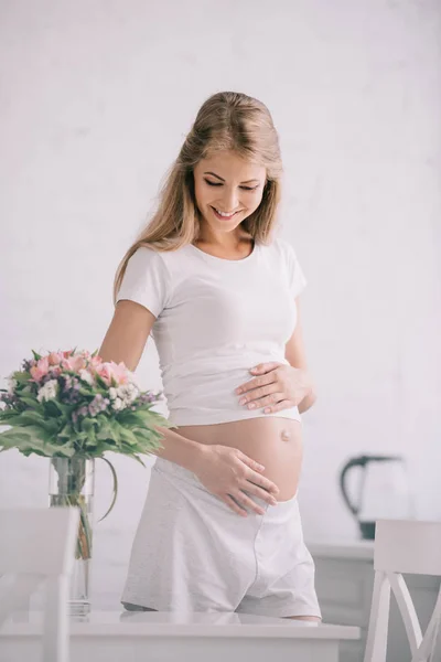 Портрет счастливой беременной женщины, стоящей за столом с букетом цветов в вазе дома — стоковое фото