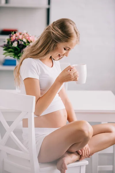 Seitenansicht einer lächelnden Schwangeren mit einer Tasse Tee, die zu Hause auf einem Stuhl sitzt — Stockfoto