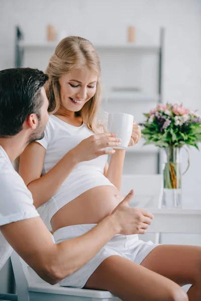 Femme enceinte souriante avec tasse de thé reposant sur une chaise avec mari à proximité à la maison — Photo de stock