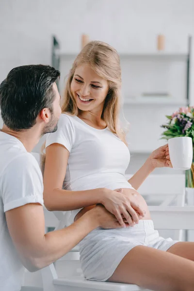 Donna incinta sorridente con una tazza di tè appoggiata sulla sedia e che tocca la pancia insieme al marito vicino a casa — Foto stock