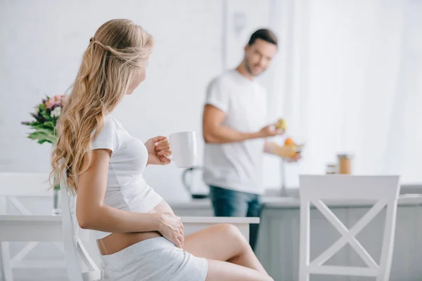 Messa a fuoco selettiva della donna incinta con tazza di tè seduta sulla sedia e marito in piedi al bancone in cucina — Foto stock