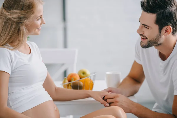 Vista laterale di donna incinta sorridente e marito che si tiene per mano a tavola a casa — Foto stock