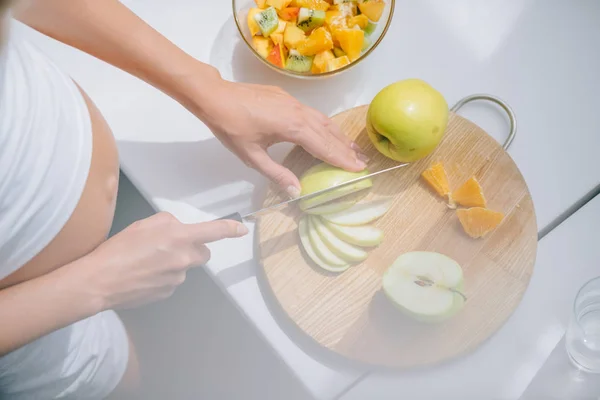Tiro recortado de mujer embarazada con cuchillo cocina frutas ensalada en casa - foto de stock