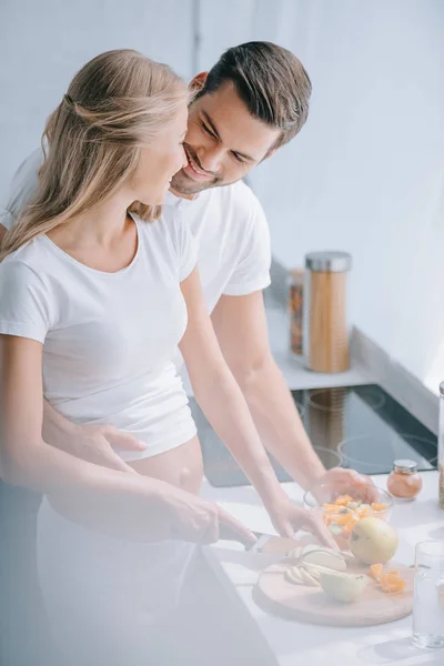 Happy pregnant woman and husband cooking fruits salad together in kitchen at home — Stock Photo