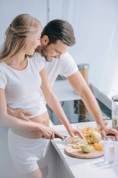 Счастливая беременная женщина и муж готовят фруктовый салат вместе на кухне дома — стоковое фото
