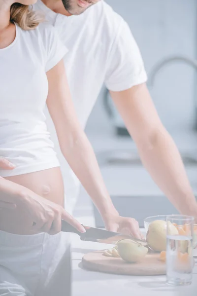 Tiro recortado de la mujer embarazada y el marido cocinar ensalada de frutas juntos en la cocina en casa - foto de stock