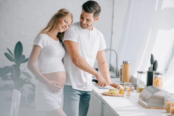 Улыбающийся мужчина режет фрукты на прилавке с беременной женой рядом на кухне дома — стоковое фото