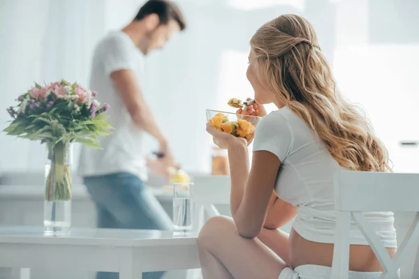 Foco seletivo da mulher grávida comendo salada de frutas enquanto o marido de pé no balcão na cozinha — Fotografia de Stock