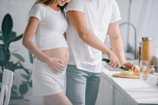 Partial view of man cutting fruits at counter with pregnant wife near by in kitchen at home — Stock Photo