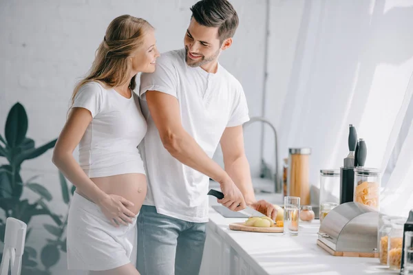 Retrato del hombre sonriente cortando frutas en el mostrador con la esposa embarazada cerca en la cocina en casa - foto de stock
