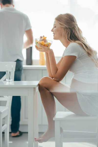 Selektiver Fokus einer lächelnden Schwangeren, die Obstsalat isst, während ihr Mann in der Küche am Tresen steht — Stockfoto