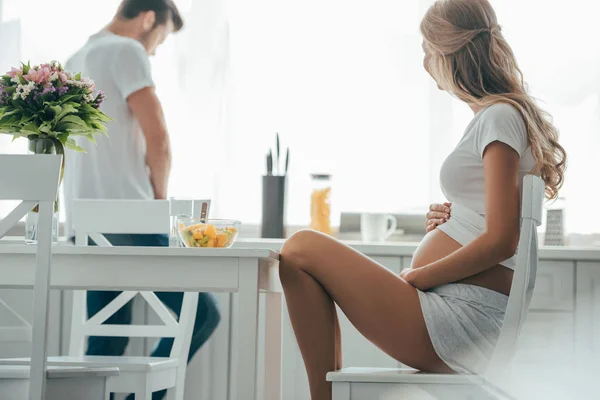 Side view of pregnant woman sitting at table with fruits salad in bowl and husband at counter in kitchen — Stock Photo