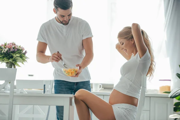 O marido trouxe a salada de frutos à esposa grávida à mesa na cozinha — Fotografia de Stock