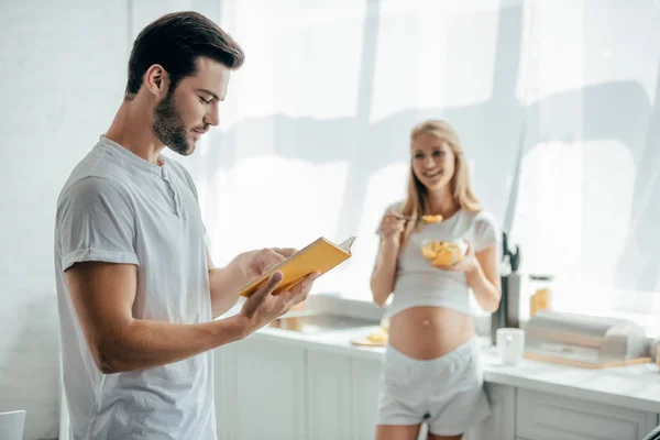 Улыбающаяся беременная жена с фруктовым салатом и муж с книгой на кухне дома — стоковое фото