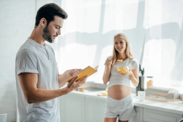 Femme enceinte souriante avec salade de fruits et mari avec livre dans la cuisine à la maison — Photo de stock