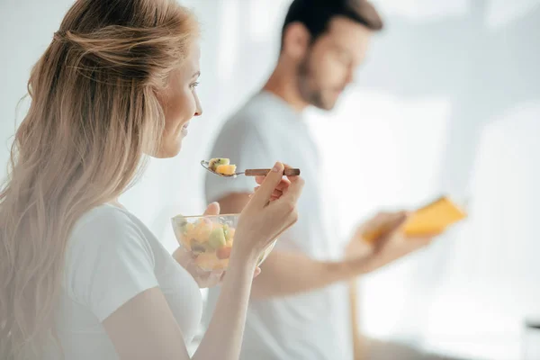 Selective focus of smiling pregnant woman eating fruits salad while husband standing at counter in kitchen — Stock Photo