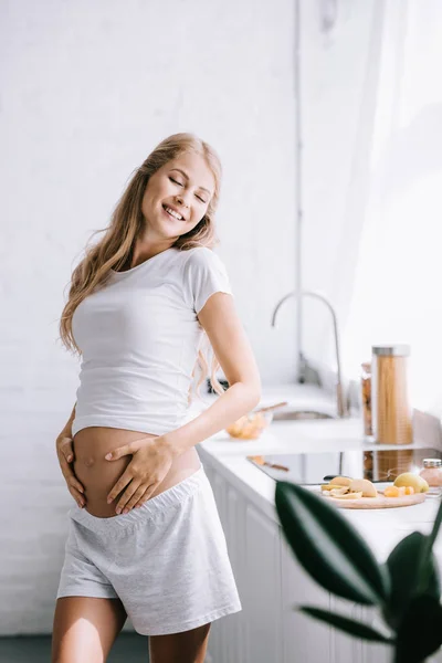 Lächelnde schöne schwangere Frau mit Händen auf dem Bauch, die zu Hause in der Küche steht — Stockfoto