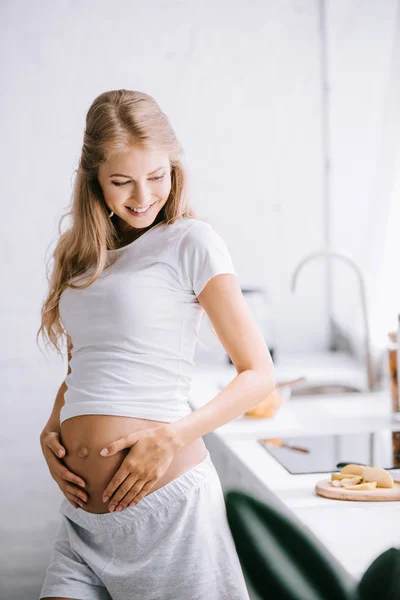 Lächelnde schöne schwangere Frau mit Händen auf dem Bauch, die zu Hause in der Küche steht — Stockfoto