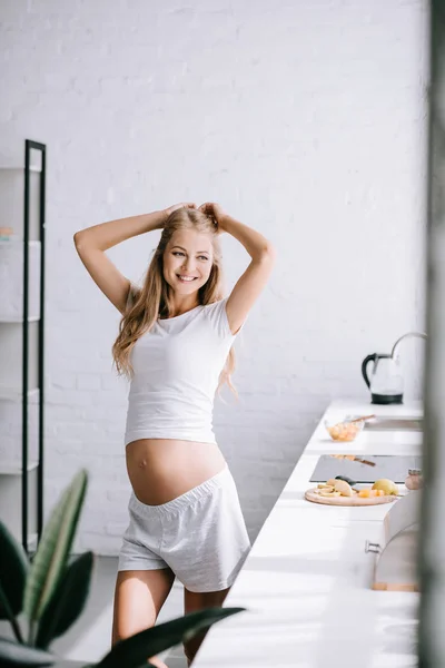 Lächelnde schöne Schwangere in weißer Kleidung, die zu Hause in der Küche steht — Stockfoto