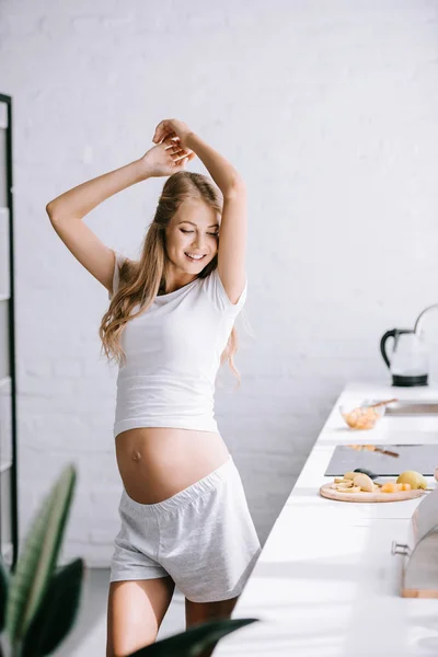 Улыбающаяся красивая беременная женщина в белой одежде, стоящая дома на кухне — стоковое фото