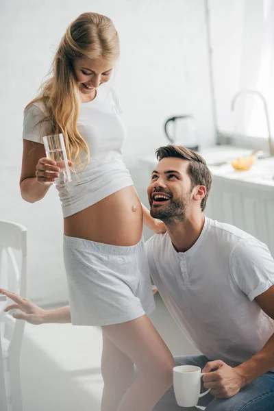Homme heureux avec tasse de café écoutant le ventre de la femme enceinte dans la cuisine à la maison — Photo de stock