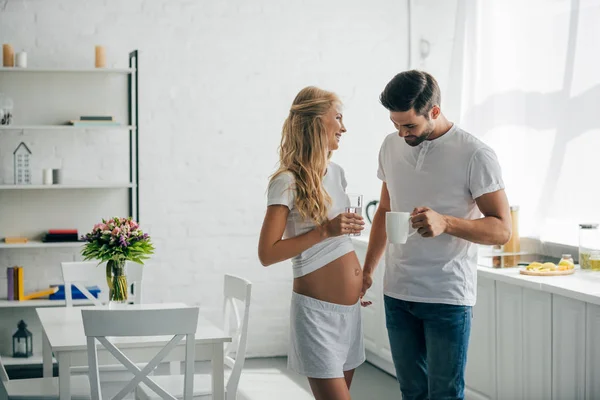 Homem com xícara de café olhando para a barriga da esposa grávida na cozinha em casa — Fotografia de Stock