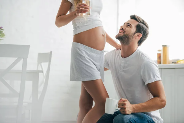 Homem com xícara de café ouvindo barriga de esposa grávida na cozinha em casa — Fotografia de Stock