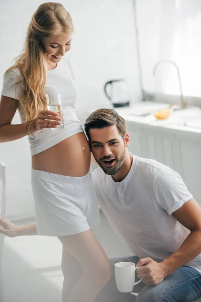 Homem com xícara de café ouvindo barriga de esposa grávida na cozinha em casa — Fotografia de Stock