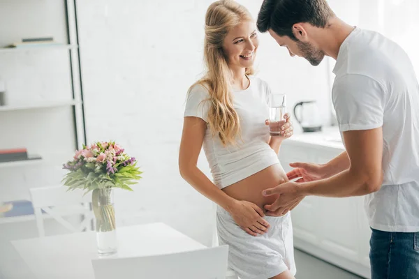 Husband touching belly of happy pregnant wife with glass of water at home — Stock Photo