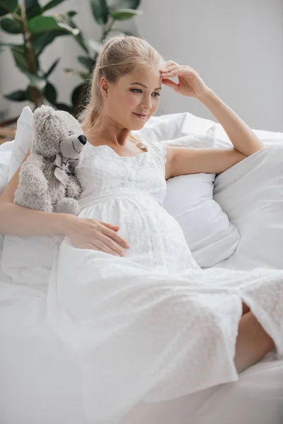 Retrato de mujer embarazada en camisón blanco con oso de peluche descansando en el sofá en casa - foto de stock