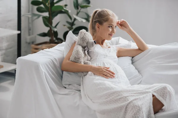 Улыбающаяся беременная женщина в белой ночнушке с плюшевым мишкой, отдыхающим дома на диване — стоковое фото