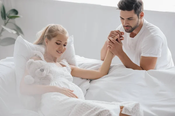 Мужчина и беременная жена в белой ночнушке на диване, держась за руки дома — стоковое фото