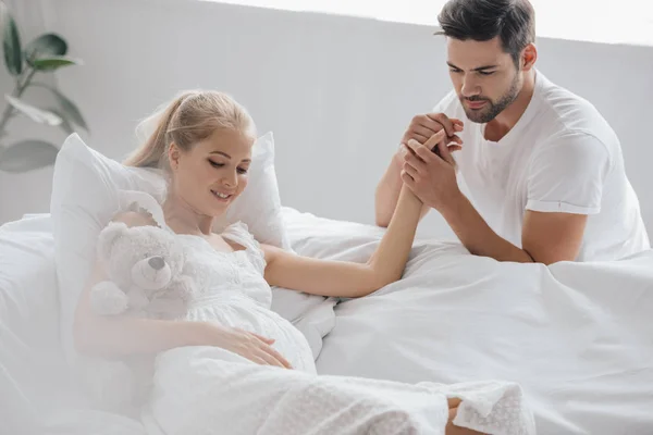 Uomo e moglie incinta in camicia da notte bianca sul divano tenendosi per mano a casa — Foto stock