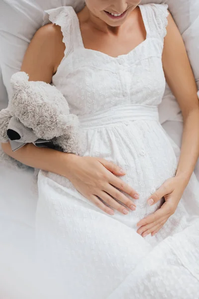 Overhead view of smiling pregnant woman in white nightie with teddy bear resting on sofa at home — Stock Photo