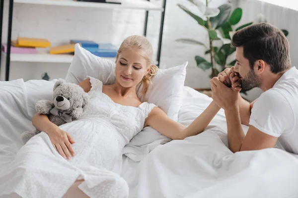 Uomo e moglie incinta in camicia da notte bianca sul divano tenendosi per mano a casa — Foto stock