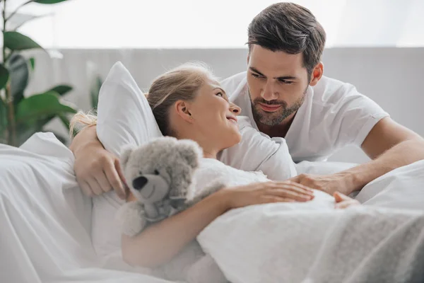 Pregnant woman in white nightie with teddy bear on sofa and husband near by at home — Stock Photo