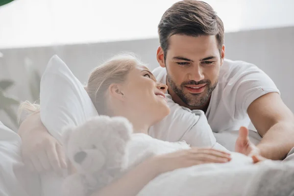 Ritratto di donna incinta in camicia da notte bianca con orsacchiotto sul divano e marito vicino a casa — Foto stock