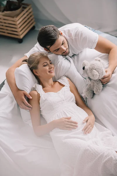 Vista de ángulo alto del hombre con oso de peluche cerca de la esposa embarazada en camisón blanco en el sofá en casa - foto de stock
