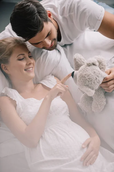 Retrato de hombre con oso de peluche y esposa embarazada sonriente en camisón blanco en el sofá en casa - foto de stock