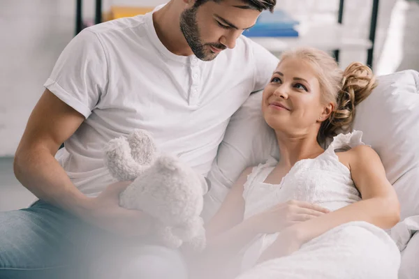 Портрет мужчины с плюшевым мишкой, сидящего рядом с беременной женой в белой ночнушке на диване дома — стоковое фото