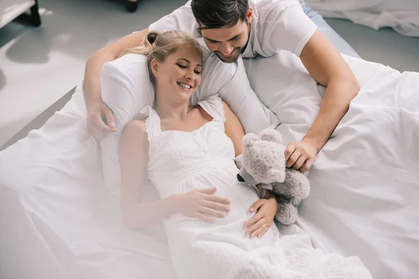 Мужчина с плюшевым мишкой возле улыбающейся беременной жены в белой ночнушке на диване дома — стоковое фото