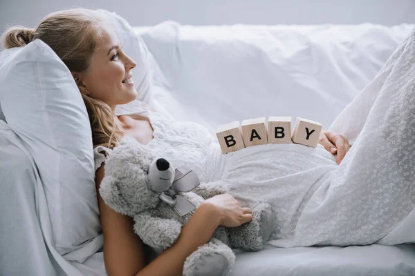 Vista lateral da mulher grávida sorridente em camisa de noite branca com ursinho de pelúcia e blocos de madeira com letras de bebê na barriga descansando no sofá — Fotografia de Stock