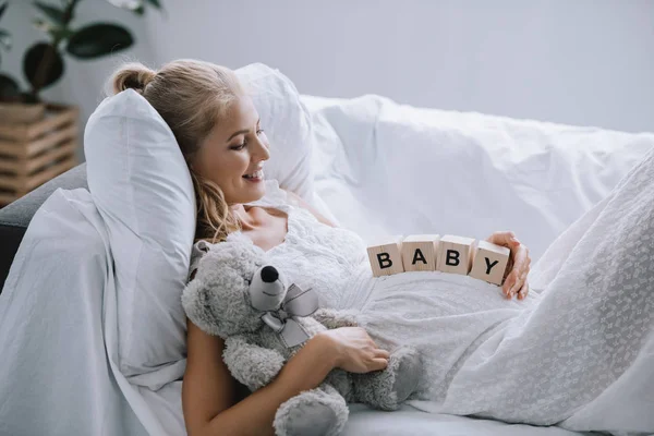 Side view of smiling pregnant woman in white nightie with teddy bear and wooden blocks with baby lettering on belly resting on sofa — Stock Photo